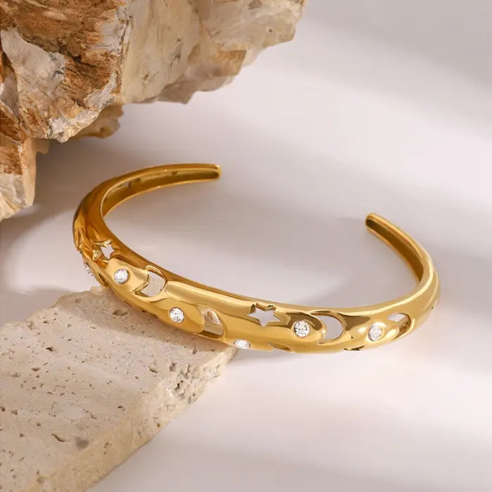 Stylish gold plated bangle with moon and star motifs, accented with sparkling stones, displayed on neutral rocks.