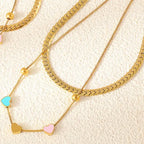 Several layers of gold chain necklaces featuring colorful heart pendants displayed on a textured surface.