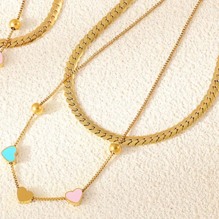 Several layers of gold chain necklaces featuring colorful heart pendants displayed on a textured surface.