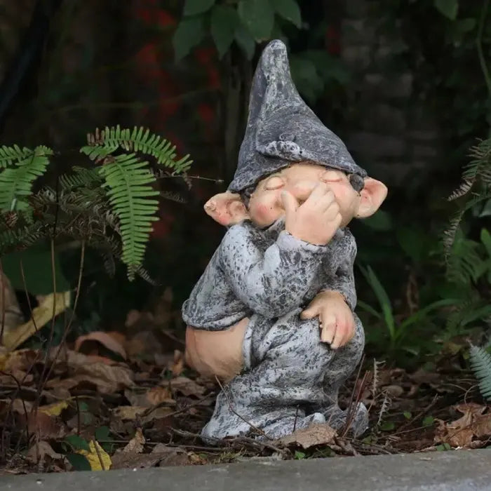 Naughty garden gnome sculpture placed in a shaded garden with leaves and plants around.