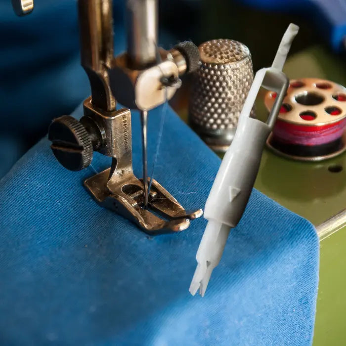Needle threader next to sewing machine foot on blue fabric with visible threading setup.