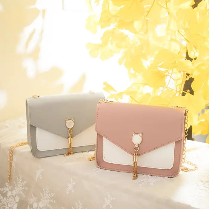 Pink and gray crossbody bags with cat charms and tassels displayed together on a lace-covered table with yellow floral decor.