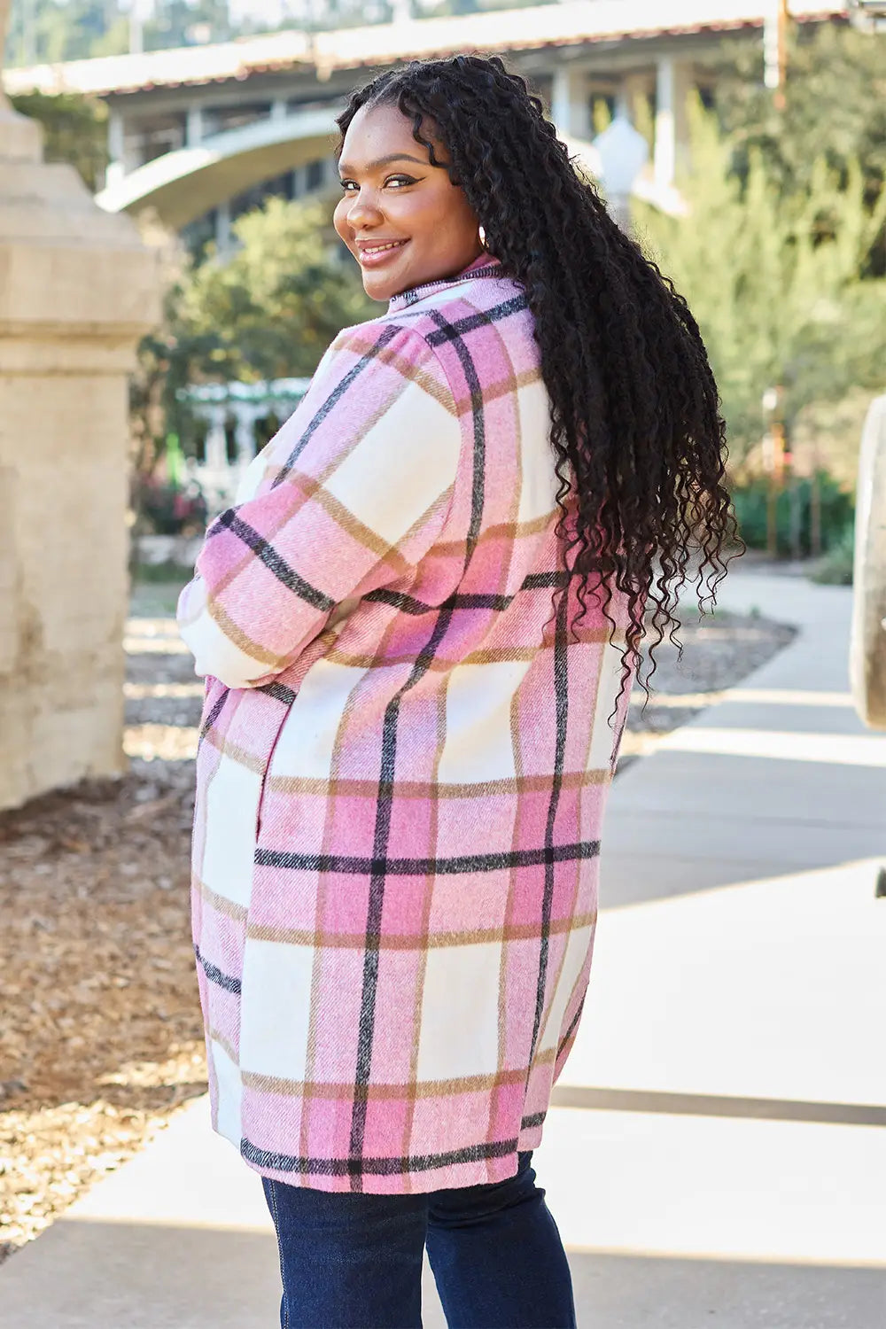 Plus-size woman wearing a pink and white plaid wool coat, styled with dark jeans, smiling outdoors near an arched bridge.