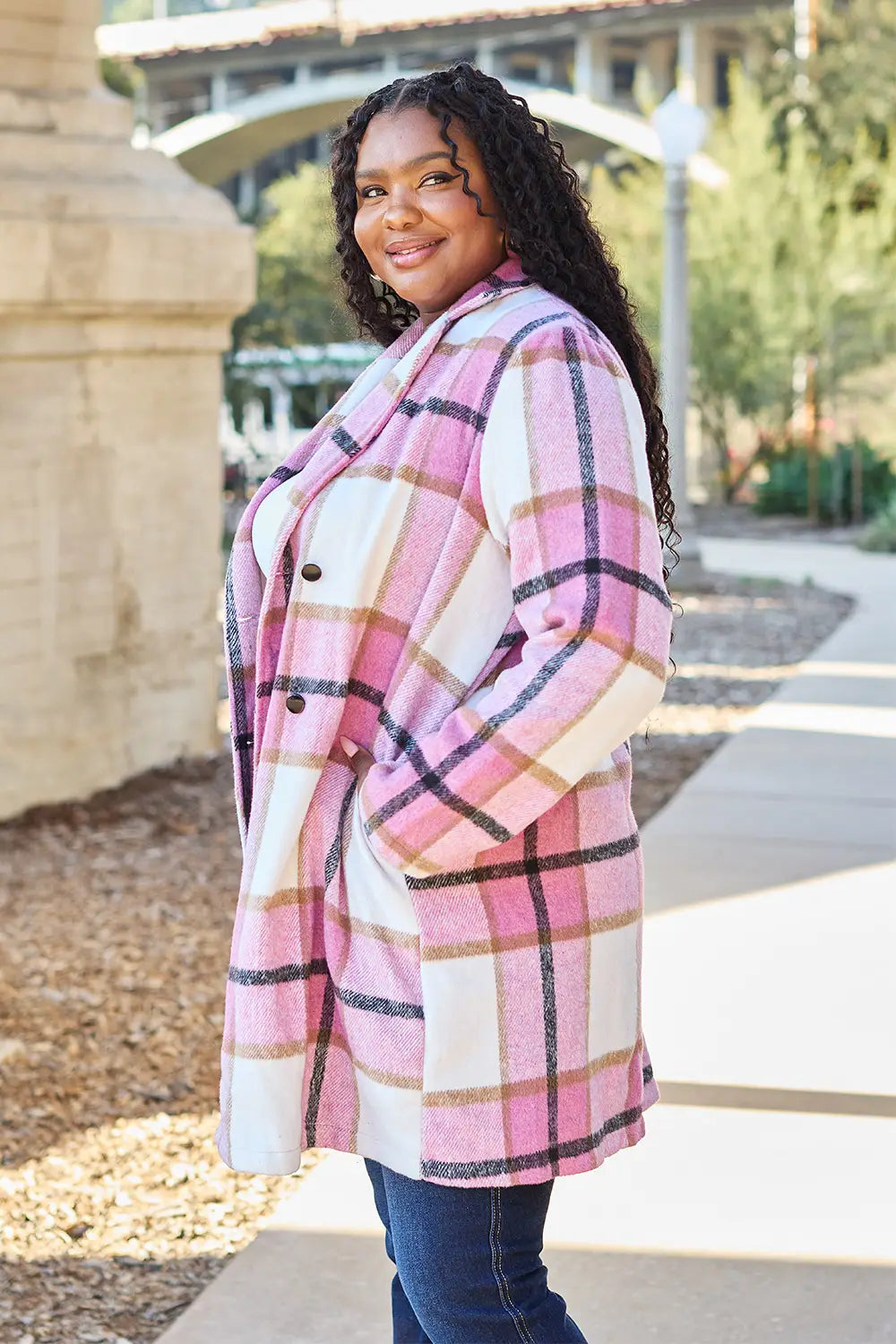 Side view of a plus-size woman wearing a pink and white plaid wool coat paired with dark blue jeans in an outdoor setting.