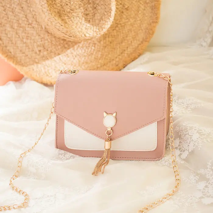 Pink crossbody bag with gold chain, cat charm, and tassel detail placed on lace fabric with a straw hat background.