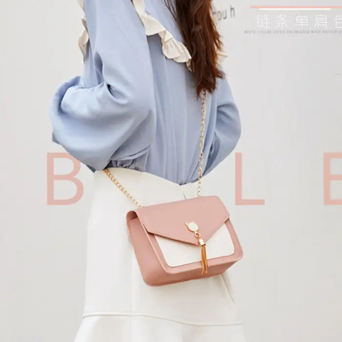 Pink crossbody bag with gold chain and cat charm shown worn by a model in a light blue blouse and white skirt.