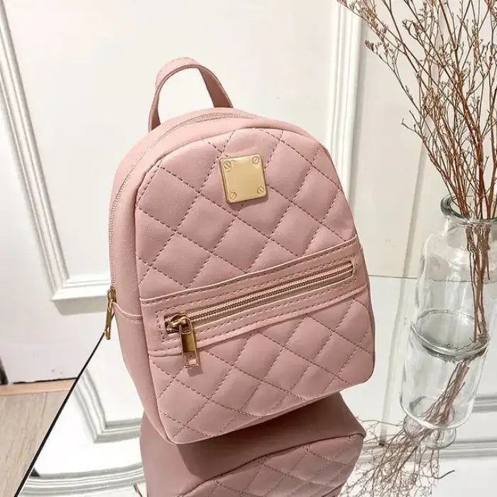 Pink quilted mini backpack with gold metal plate and front zipper pocket, displayed on reflective surface next to glass vase