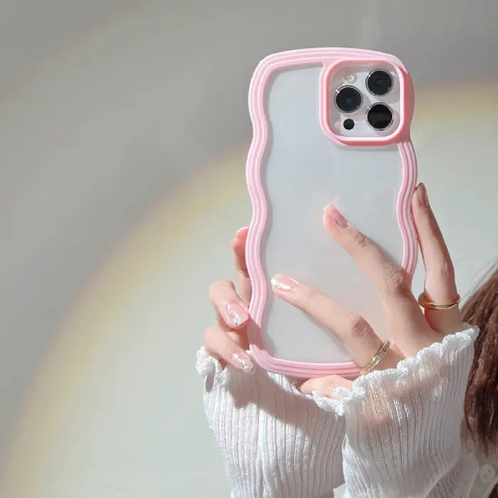 A pink wavy-border iPhone case held by a hand with soft pink nails and gold rings.