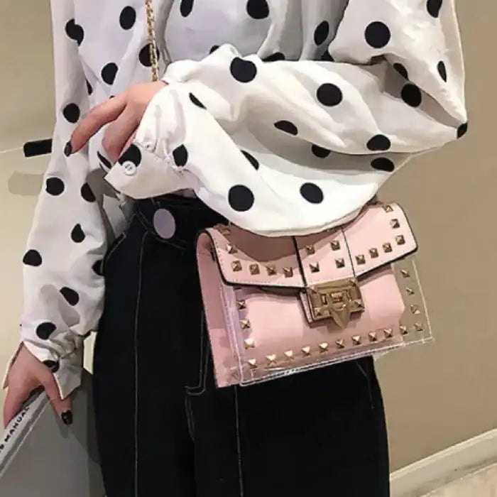 Person wearing white polka dot blouse and black pants, carrying small pink studded handbag with gold hardware