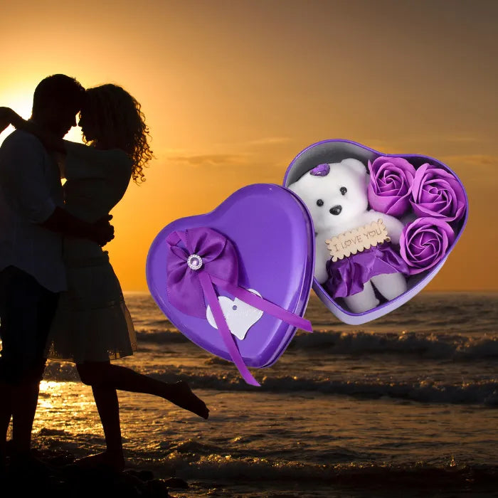 Purple heart-shaped gift box with teddy bear and roses overlaid on romantic sunset beach silhouette