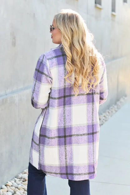 Back view of a woman with blonde curls wearing a purple plaid wool coat paired with dark jeans in a minimalist outdoor setting.