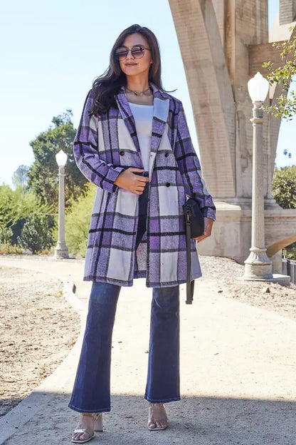 Full view of a woman in a double-breasted purple plaid wool coat paired with flared jeans and heels under an arched bridge.