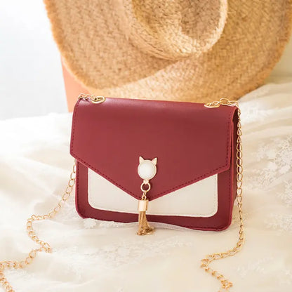 Red crossbody bag with gold chain, cat charm, and tassel detail placed on lace fabric with a straw hat in the background.