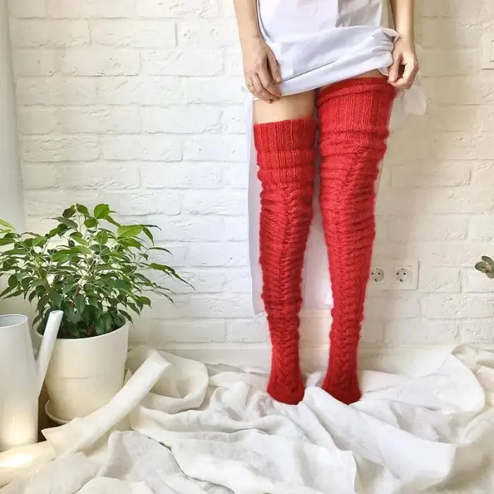 Person wearing long red knit socks, white shirt, against white brick wall with potted plant and fabric