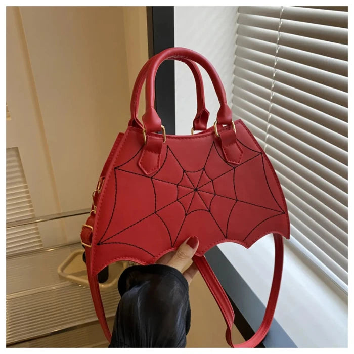 Red handbag with black spiderweb design, bat-wing shape, held by hand with red nails against white blinds background