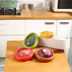 reusable fruit and vegetable storage containers with color-coded lids holding avocado, tomato, onion and lemon on wooden counter