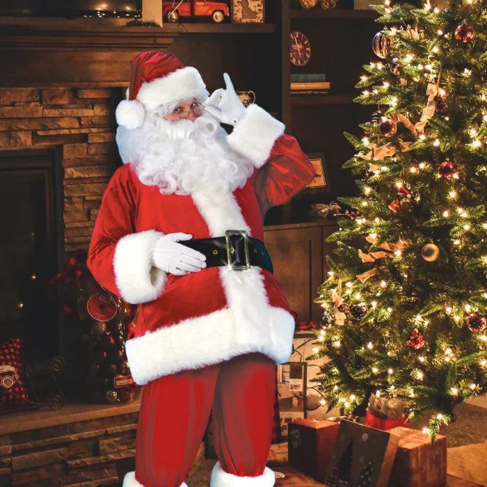 Santa costume adult posing beside a decorated Christmas tree and fireplace with wrapped presents.