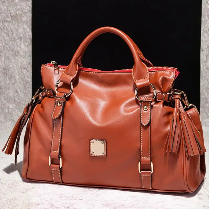 Satchel handbag in brown leather with tassel accents shown on neutral textured background