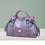Satchel handbag in light purple leather with tassels displayed on tabletop against green wall