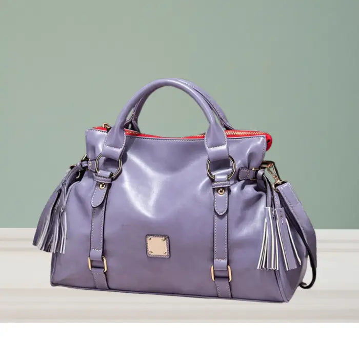 Satchel handbag in light purple leather with tassels displayed on tabletop against green wall
