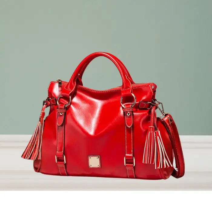 Satchel handbag in glossy red leather with tassels displayed on tabletop and green backdrop