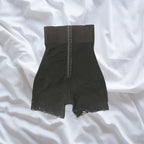 Full front view of black shapewear with high waistband and lace leg trim on white bedding.