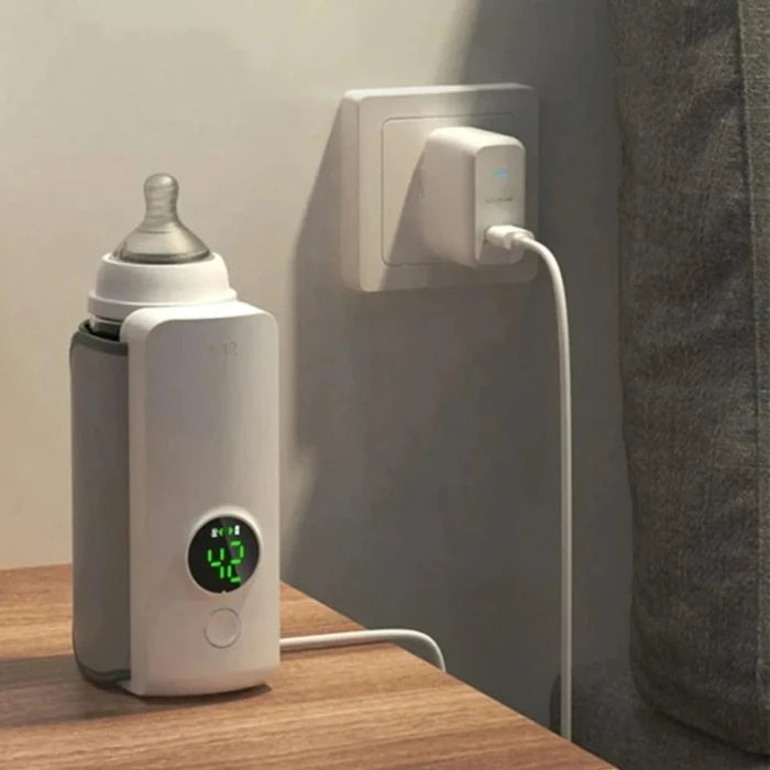 Smart bottle warmer with digital display plugged into wall outlet, baby bottle inside, on wooden surface near gray couch