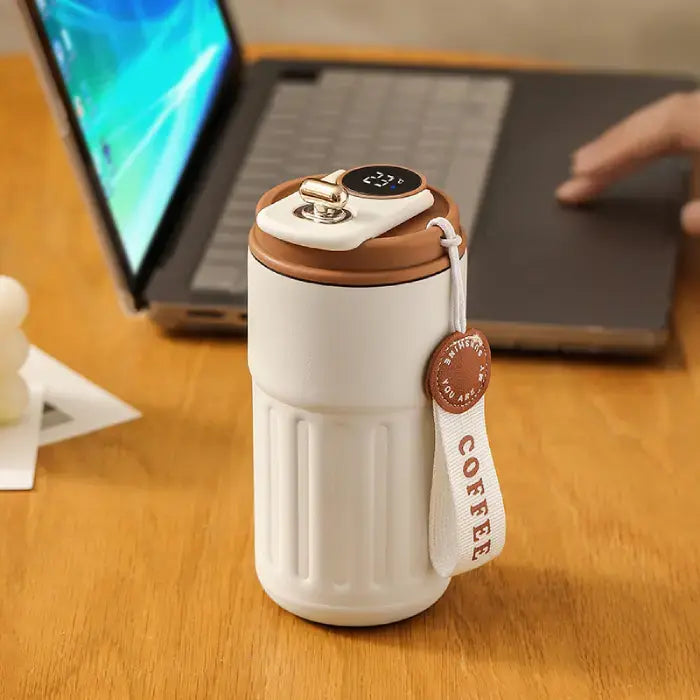 White smart coffee mug with digital display on wooden desk next to laptop, showcasing modern workspace