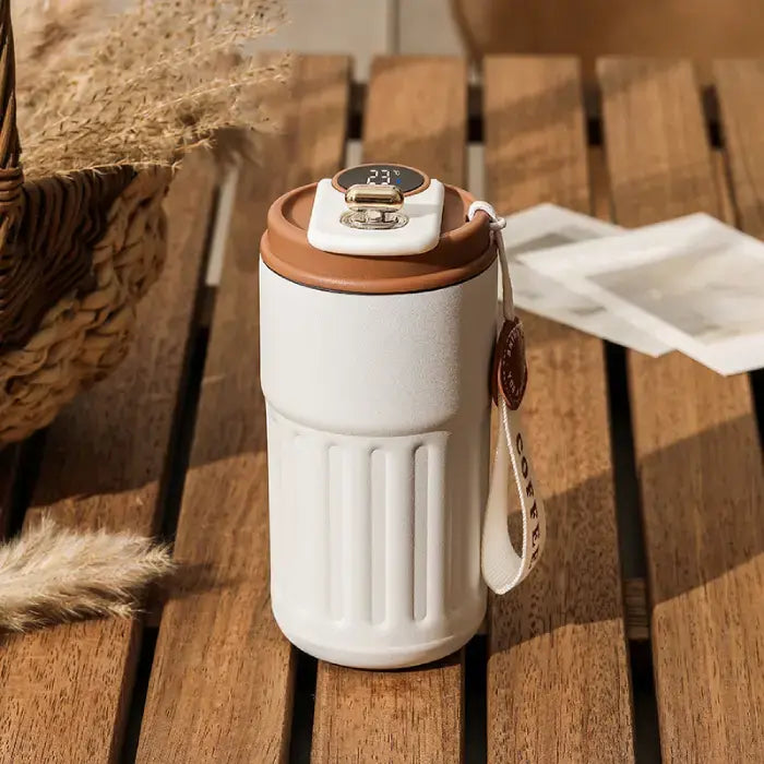 White smart thermos with digital temperature display on wooden surface, surrounded by wheat stems and woven basket