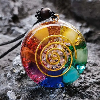 Closeup of chakra healing pendant featuring gold spiral and rainbow stones on a rocky surface.