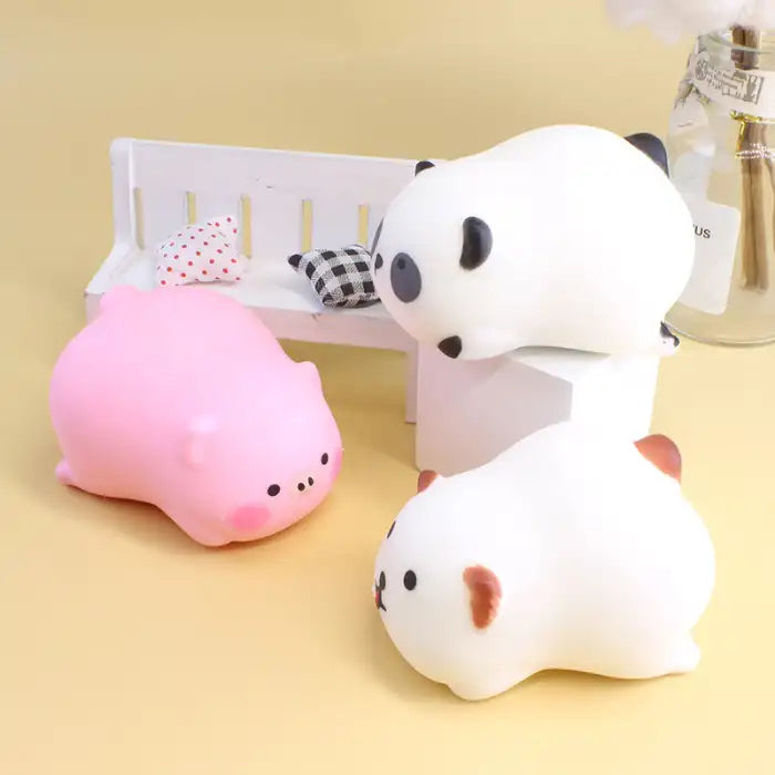 Squeeze toys of panda, pig, and bear arranged in playful scene with bench and pillows.