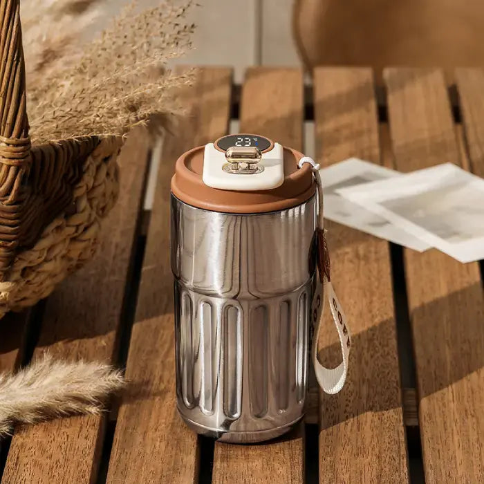 Stainless steel smart coffee mug with digital display on wooden surface, surrounded by natural elements