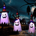 String lights with glowing ghost faces and witch hats hung outdoors near barn in spooky Halloween setup.