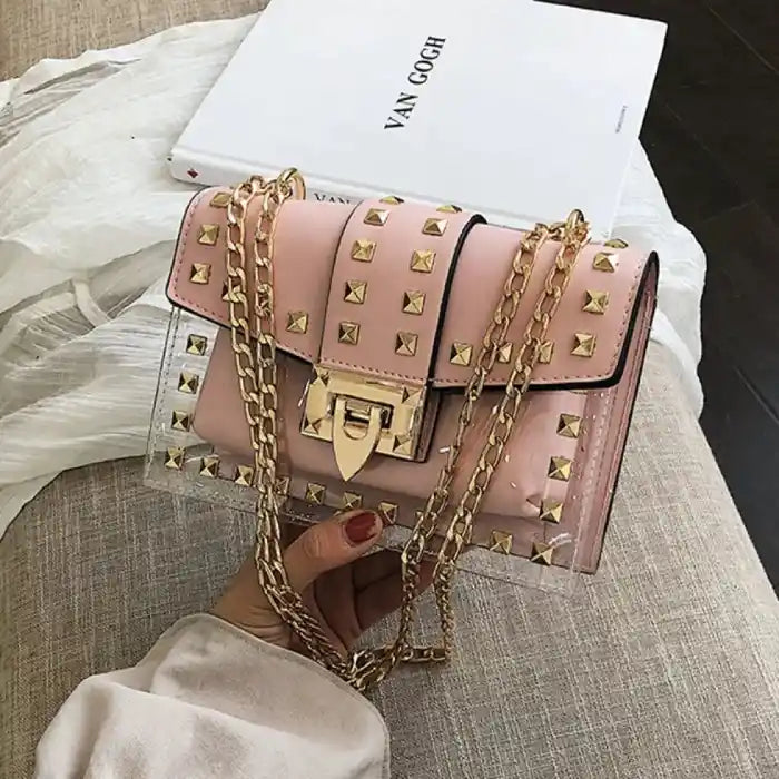 Close-up of pink leather purse with gold studs, chain strap, and lock closure, displayed on textured surface near white book