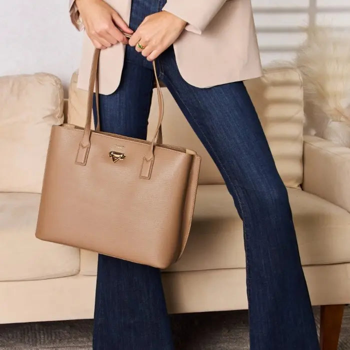 Close-up of tan leather tote bag held against blue jeans and beige blazer, showcasing bag's size and style