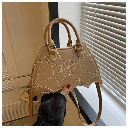 Tan handbag with white spiderweb stitching, bat-wing shaped, held by hand with red nails against white window blinds