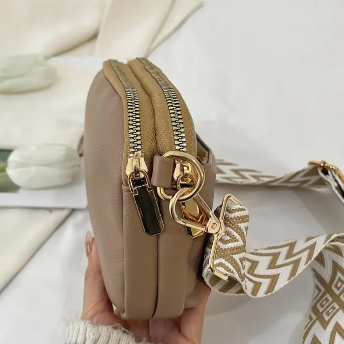 Taupe crossbody bag with gold-tone zippers and a chevron strap, showcasing a sleek and compact design.