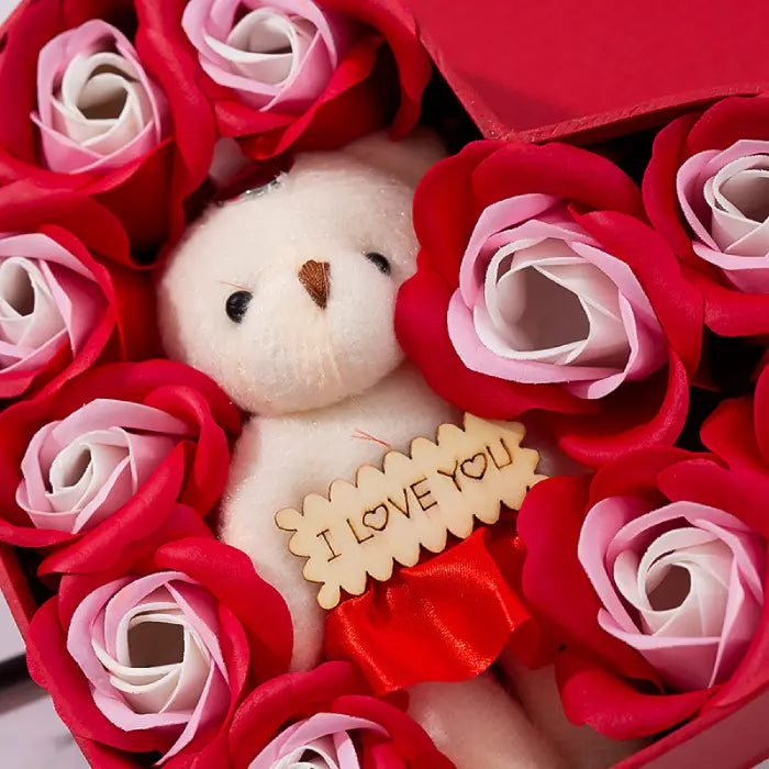 Close-up of teddy bear surrounded by red and pink artificial roses in a heart-shaped gift box.