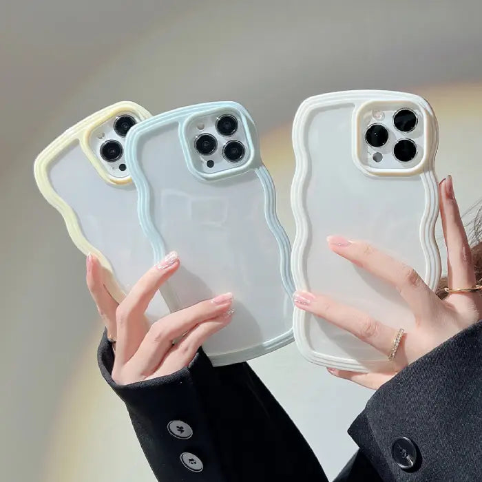 Three wavy-frame iPhone cases in pastel yellow, blue, and white held by a person wearing a black coat.