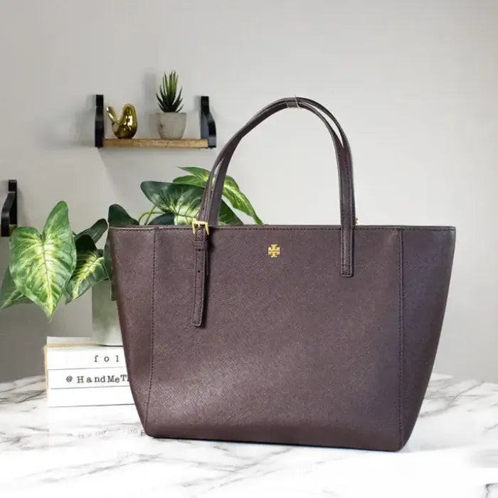 Tory Burch Emerson brown tote on marble table, elegantly styled with plants, perfect for chic settings.