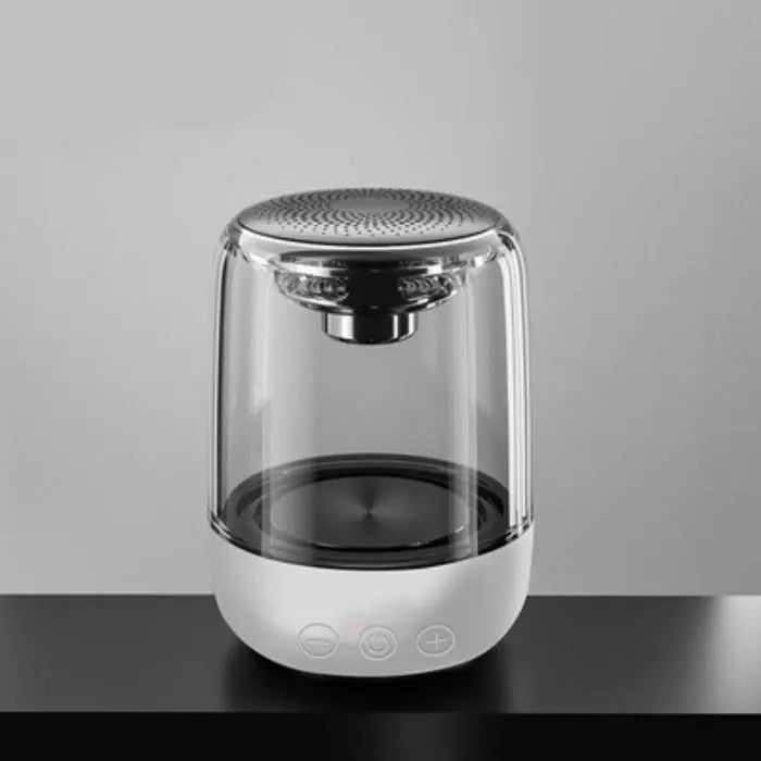 Wireless speaker with clear glass body, silver top, white base with control buttons, placed on a dark surface