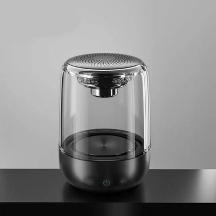 Transparent wireless speaker with silver perforated top, glass body, and black base on a dark surface