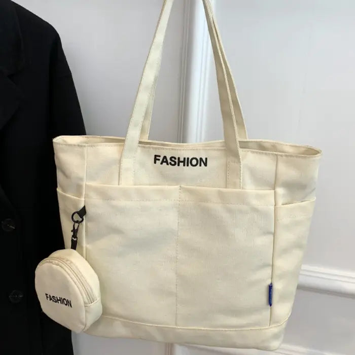 White canvas tote bag with mini pouch labeled "FASHION," hanging on a wall for a minimalist display.