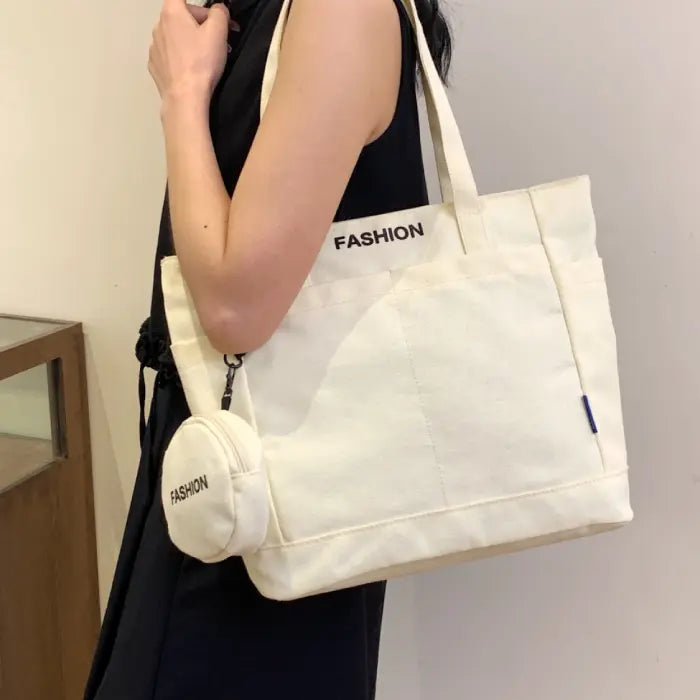 White canvas tote bag with mini pouch labeled "FASHION," carried by a woman in a black sleeveless dress.