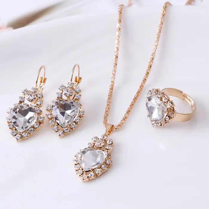 White gemstone heart jewelry set with gold necklace, earrings, and ring surrounded by sparkling crystals.