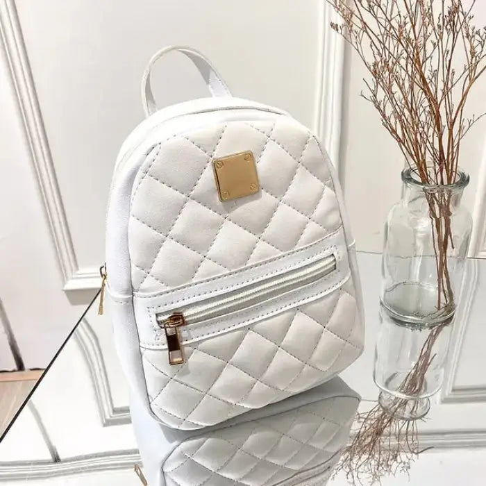 White quilted mini backpack with gold metal plate and front zipper pocket, displayed on reflective surface next to glass vase