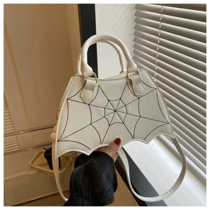 White handbag with black spiderweb stitching, bat-wing shaped, held by hand with red nails against white blinds