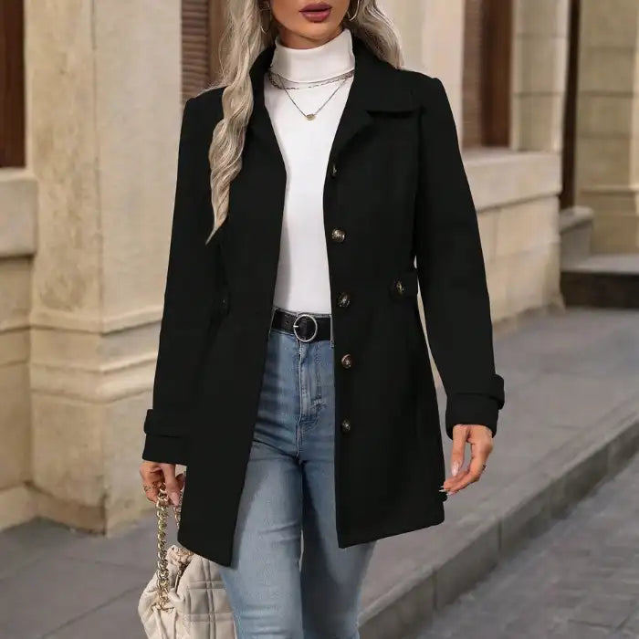 Woman wearing a black coat, white turtleneck, and blue jeans standing on a street.
