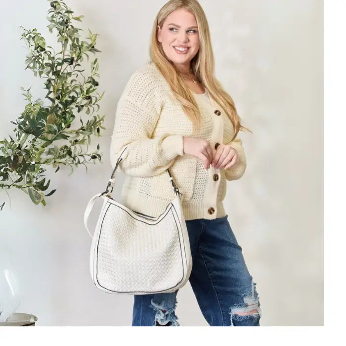 Woman in cream sweater and distressed jeans with stylish white handbag, standing beside a plant.