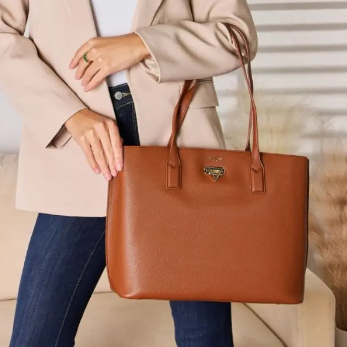 Woman holding brown leather tote bag, wearing beige coat and blue jeans, showcasing bag's size and style